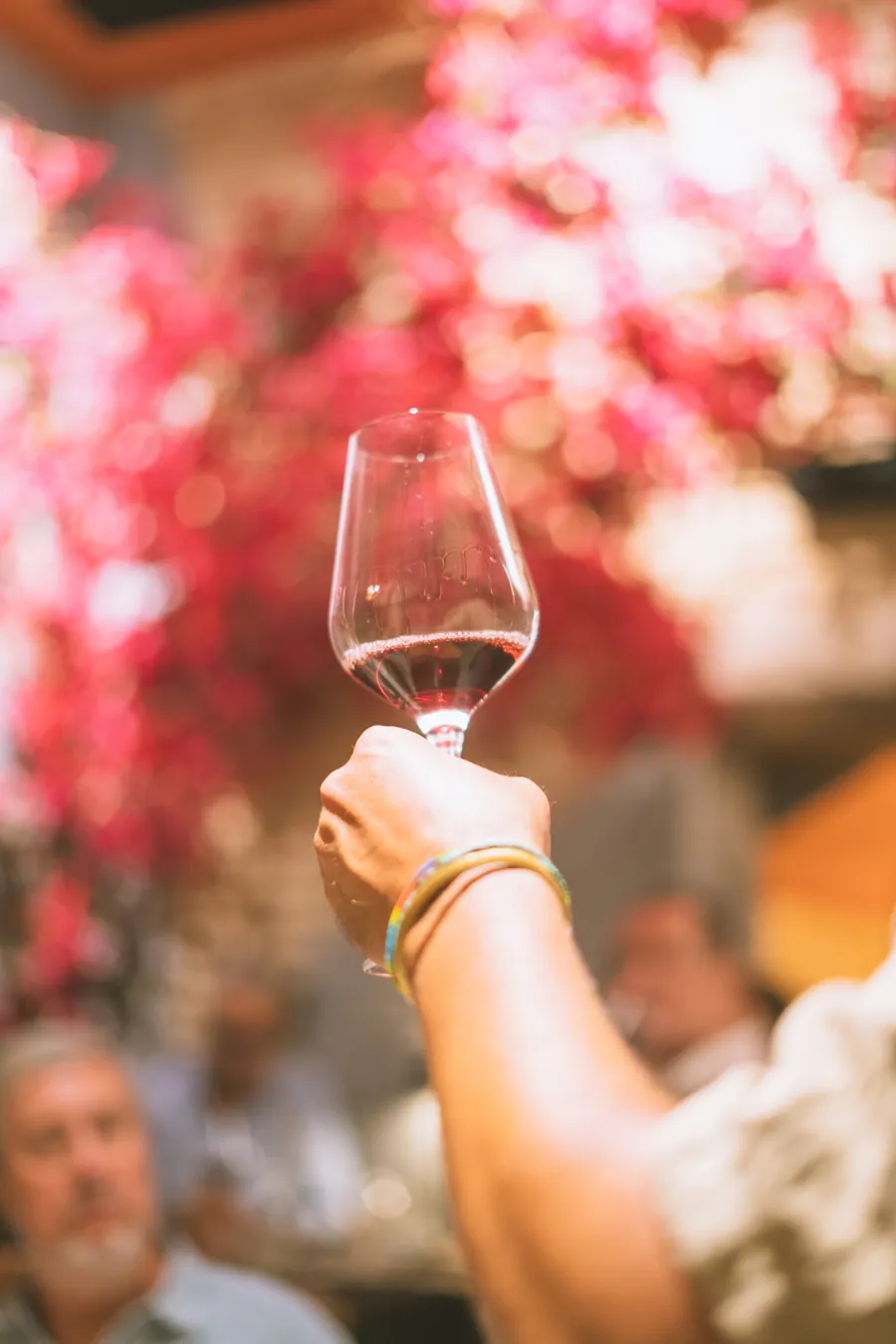 Una mano levanta una copa de vino rosado frente a un fondo de buganvillas