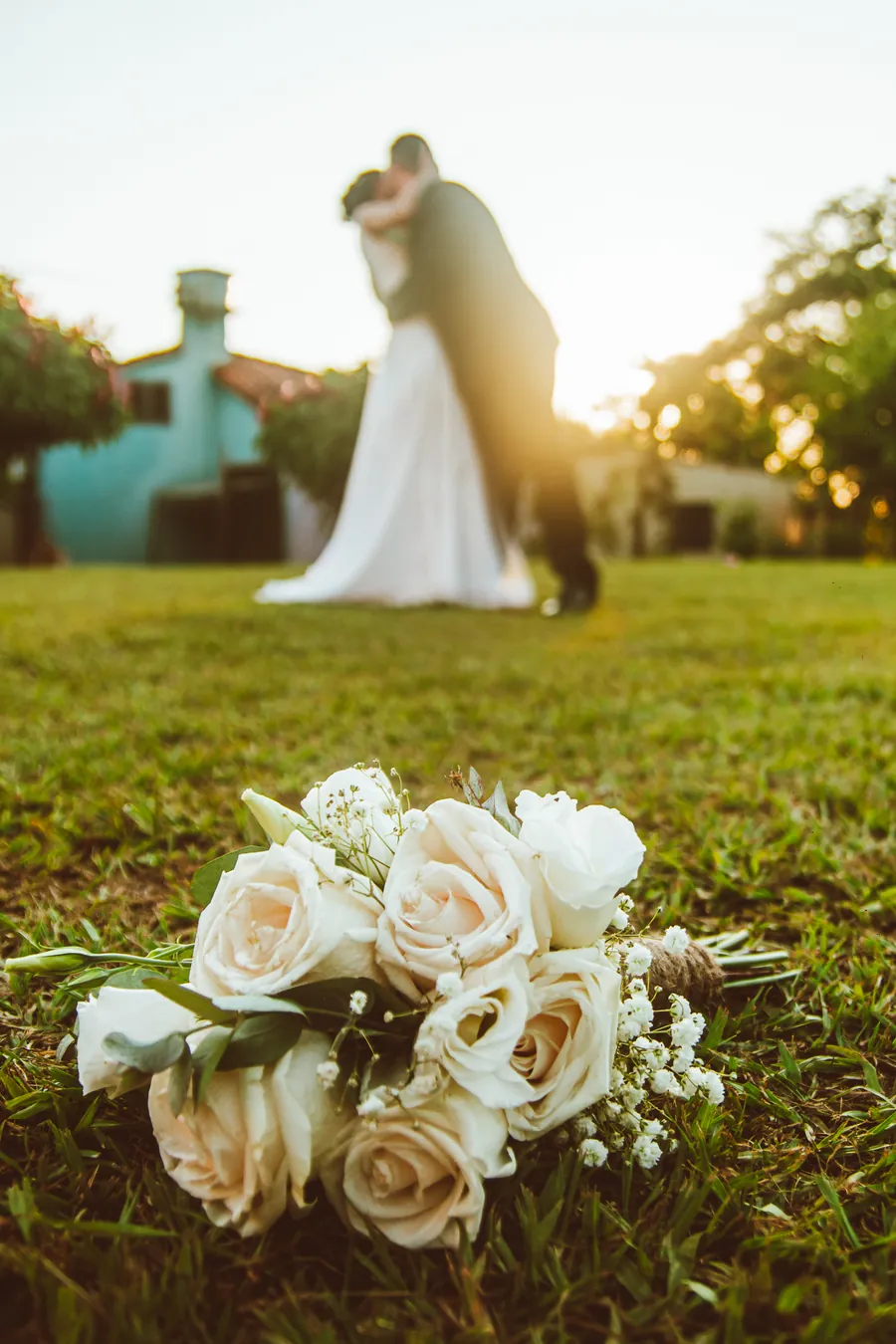 Un ramo de rosas en el césped y, al fondo desenfocado, los novios besándose