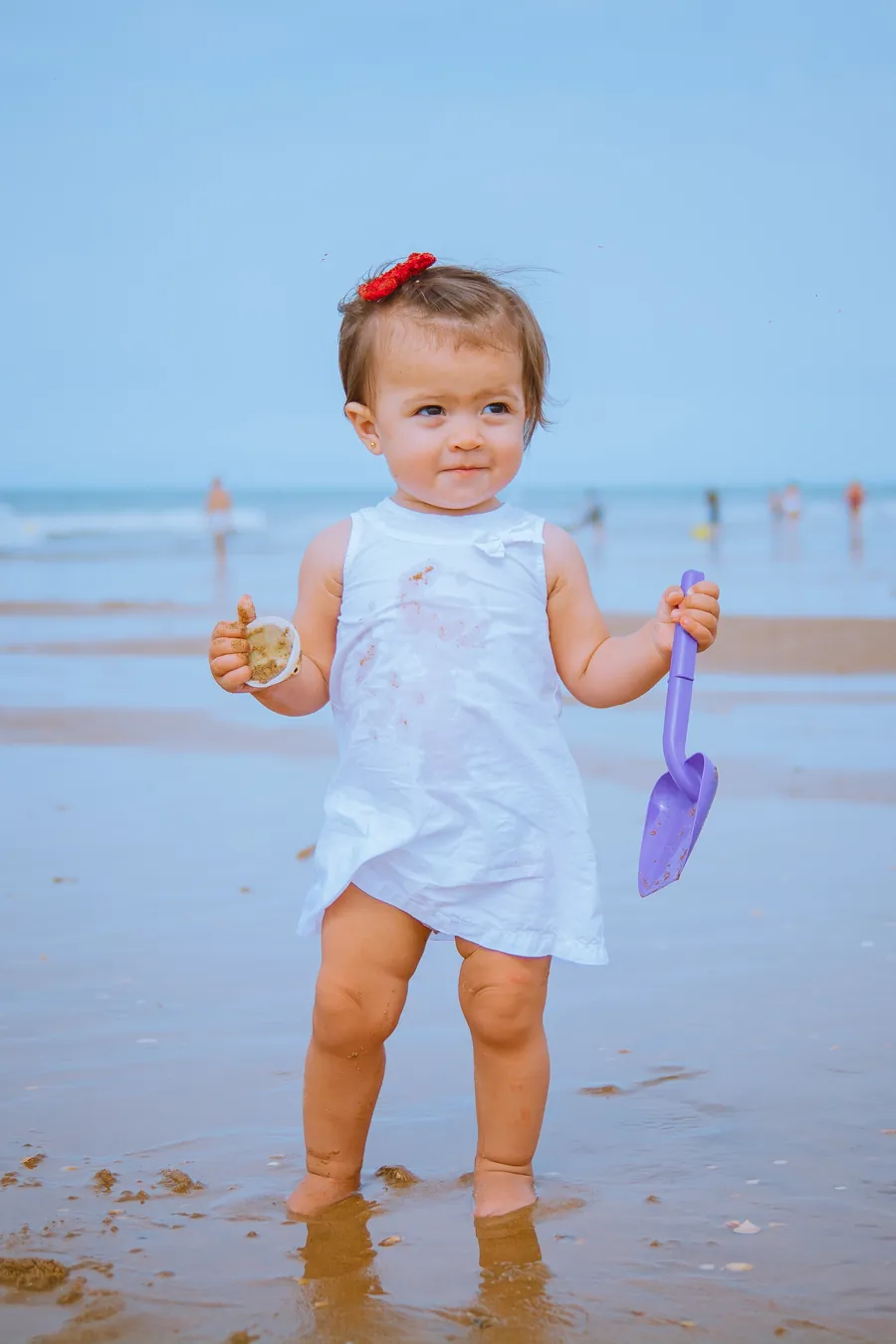 Una niña pequeña con vestido blanco y una pala en la orilla del mar